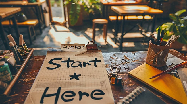 A cluttered desk with a handwritten "Start Here" sign amid scattered supplies, representing the simple act of choosing a starting point when organizing feels overwhelming