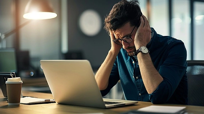Stressed man sitting at a desk in front of his computer holding his head from feeling overworked
